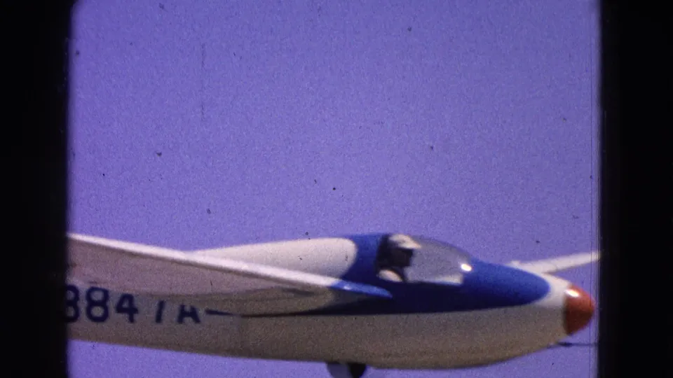 A close 8mm frame shows the glider fuselage, blue nose, cockpit, wing, and partial registration markings as it passes by.