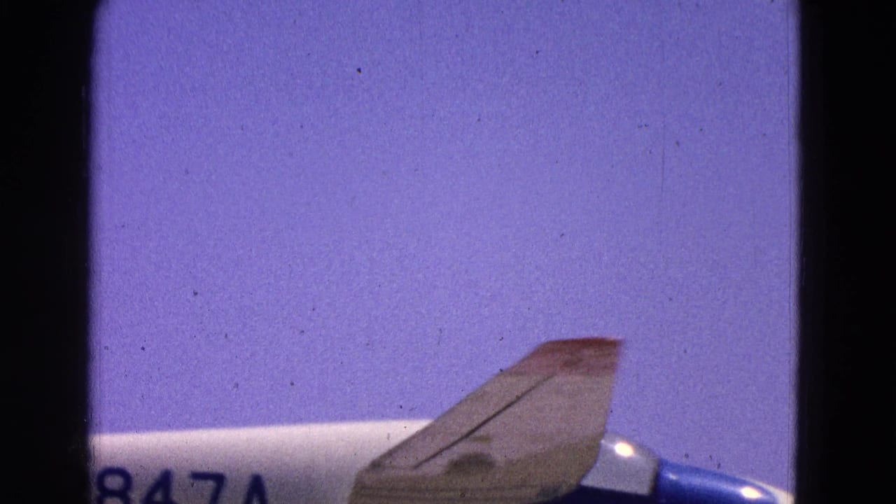 Partial blue and white airplane against a pale purple sky with film gate edges.