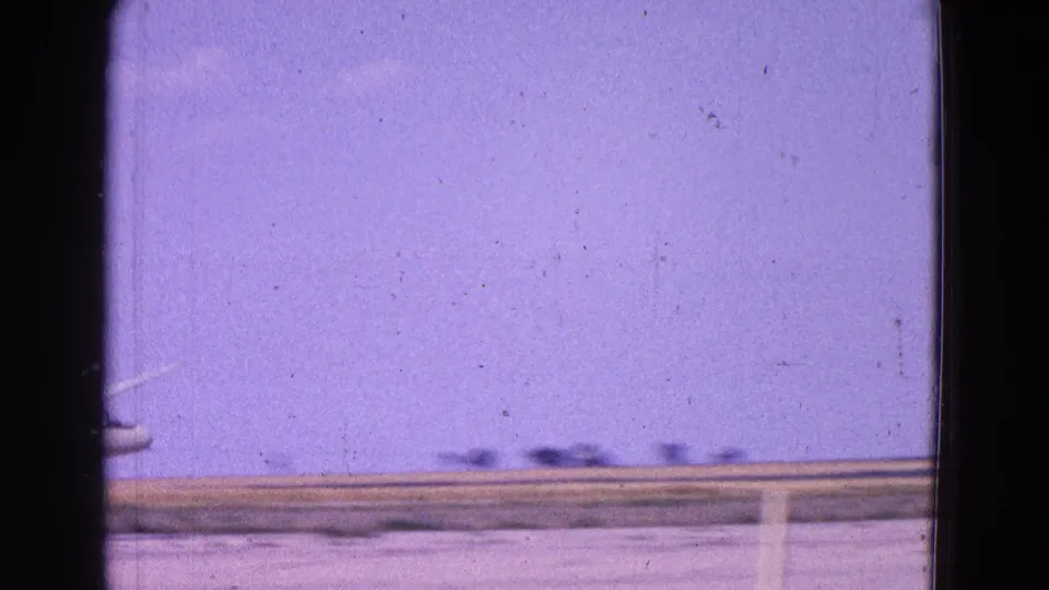 1964 Washington, DC area 8mm frame with a small white plane low over a flat landscape under a hazy purple sky.