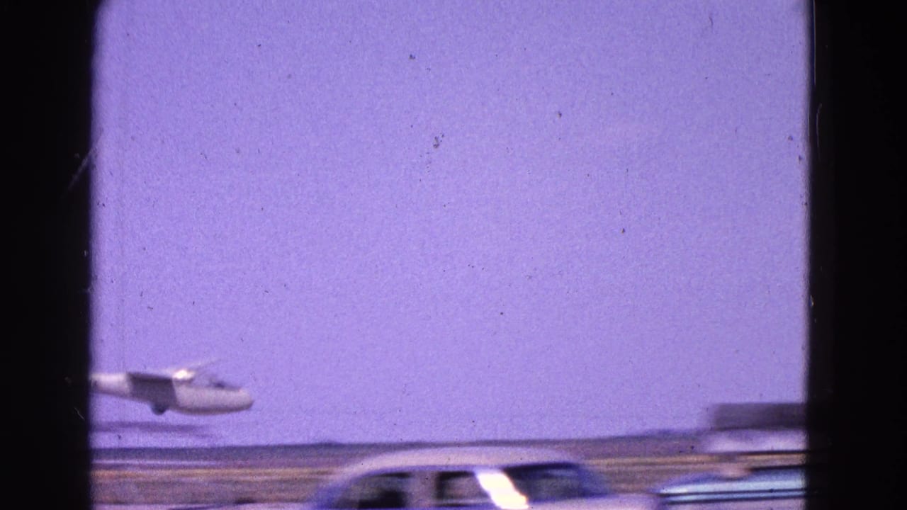 Small plane approaching over a flat landscape with a car passing in the foreground.