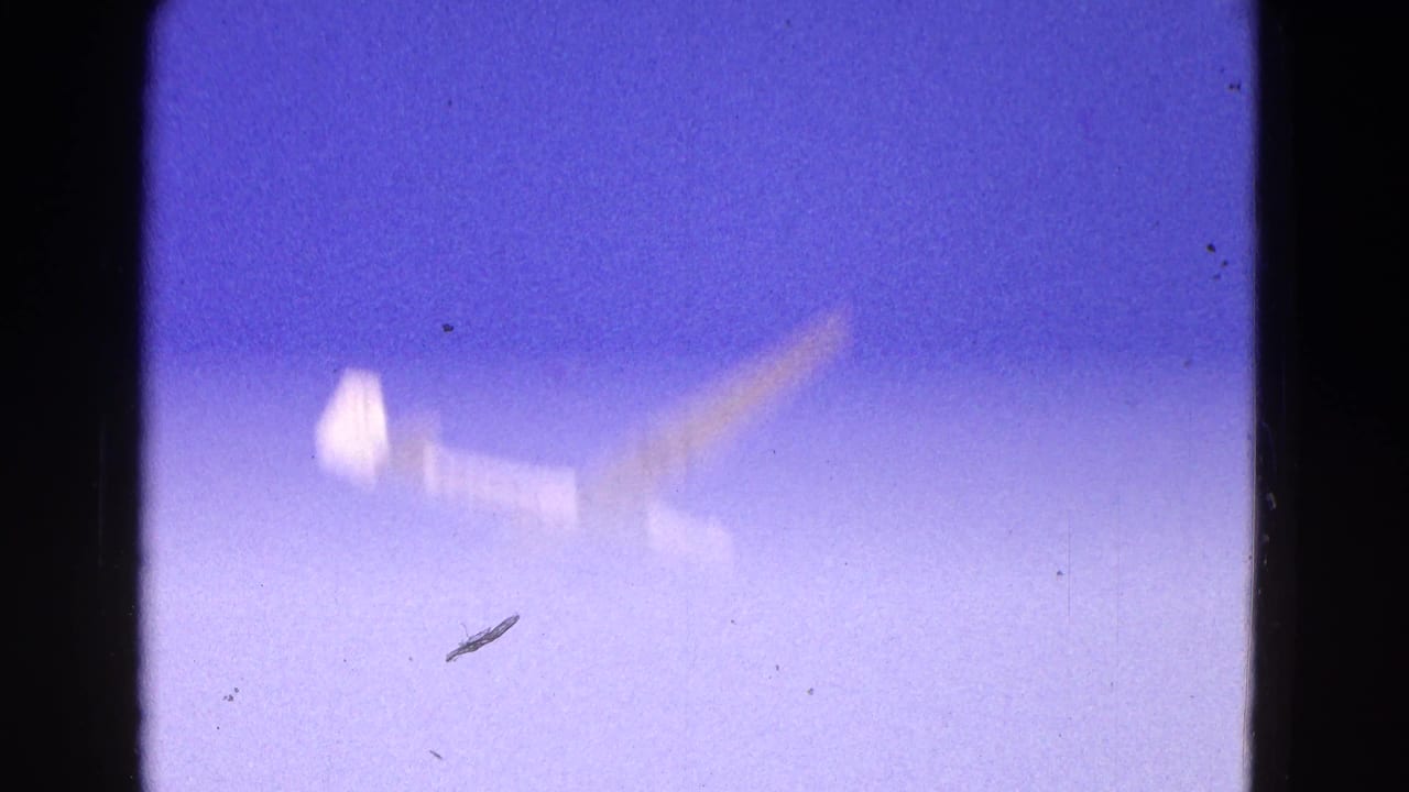 1964 8mm frame of a white glider appearing soft and distant against a purple-blue sky, with visible film grain and gate edges.