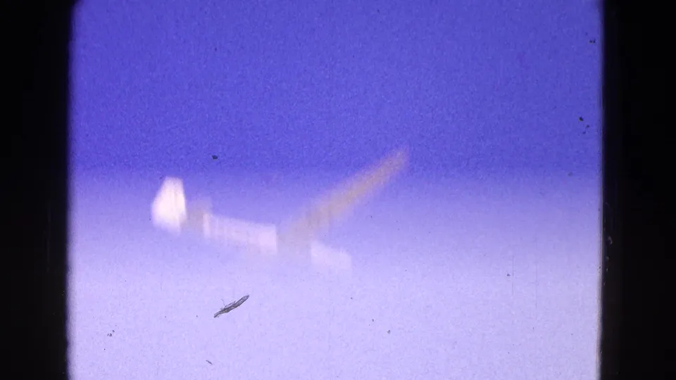1964 8mm frame of a white glider appearing soft and distant against a purple-blue sky, with visible film grain and gate edges.