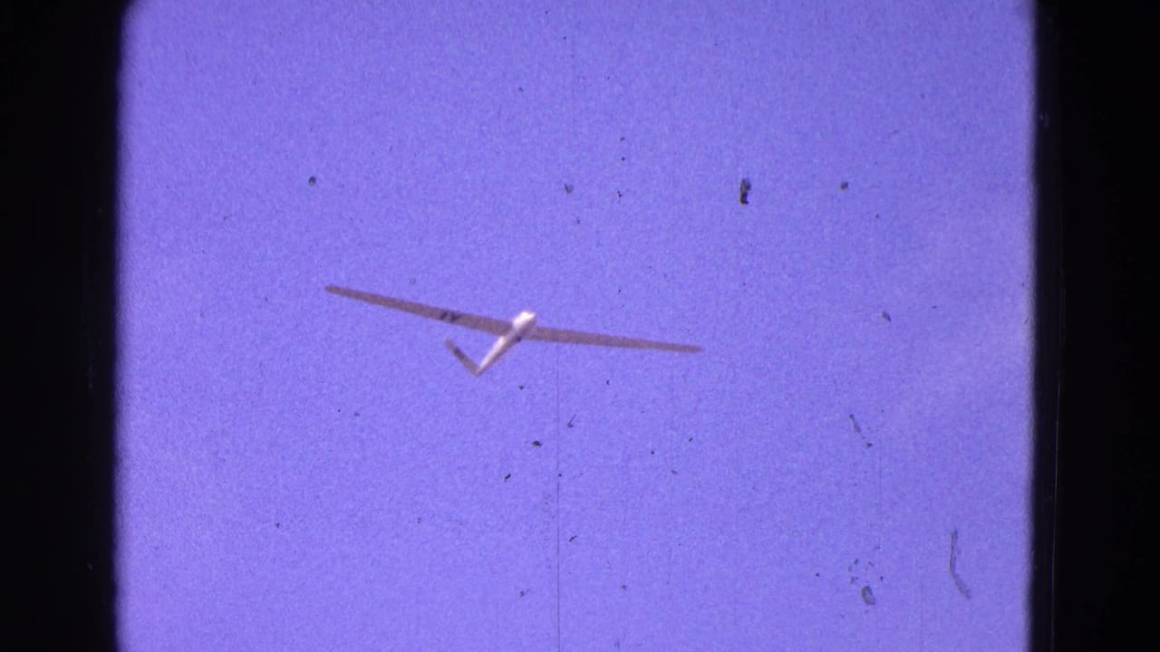 White glider viewed from below in 1964 home-movie footage, framed by dark 8mm gate edges and scattered dust marks.
