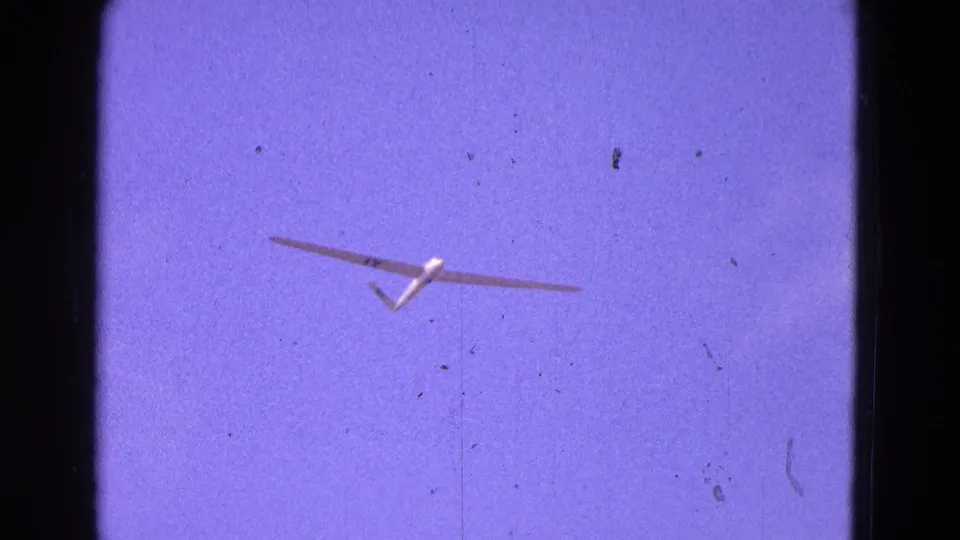 White glider viewed from below in 1964 home-movie footage, framed by dark 8mm gate edges and scattered dust marks.