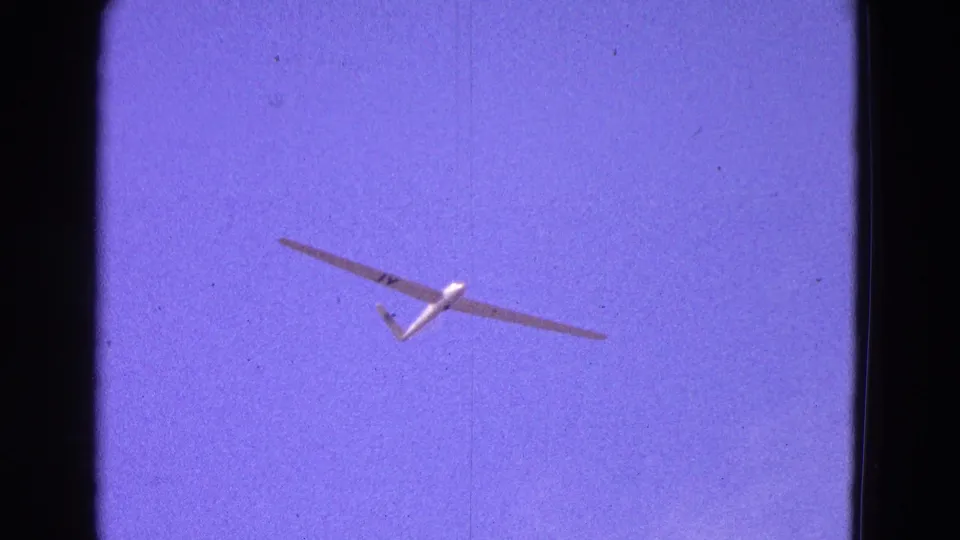 A white glider banks slightly across the purple-blue sky in 1964 8mm footage, with grain, scratches, and edge shadowing visible.