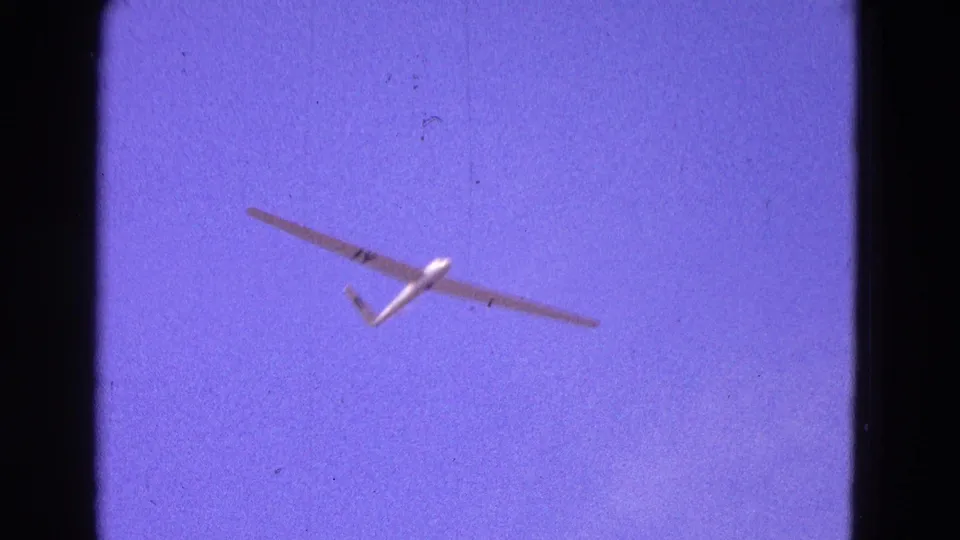 White glider centered against a purple-blue sky in 1964 Washington, DC-area home-movie footage, with 8mm grain and dust visible.