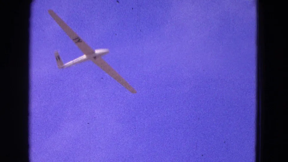 A below-view frame of the white glider, with long wings and tail markings visible in purple-tinted 1964 home-movie footage.