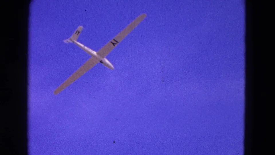 1964 8mm home-movie frame of a white glider banking through blue sky, with long wings and dark registration-style markings visible.