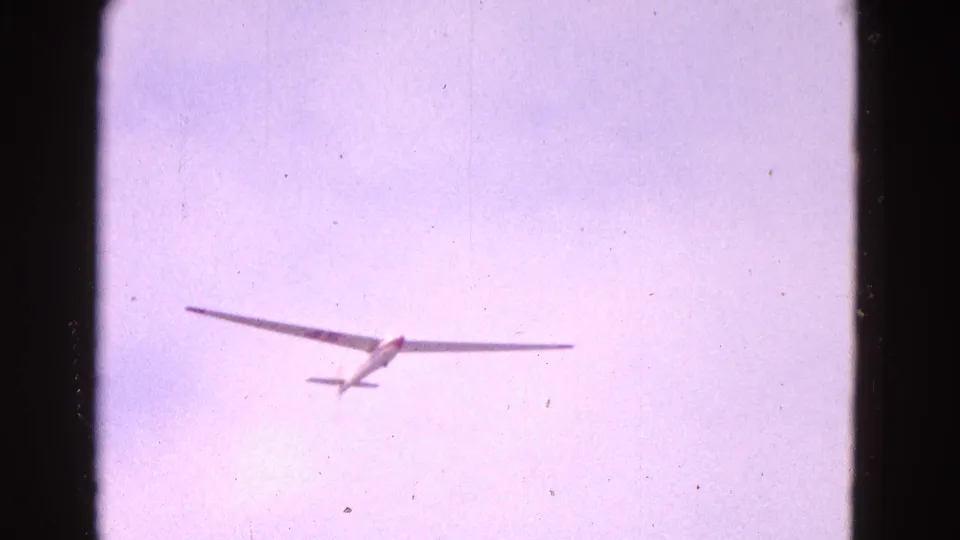 A distant white glider appears from below in pale sky, with its wings level and tail visible in this 1964 Washington DC home-movie frame.
