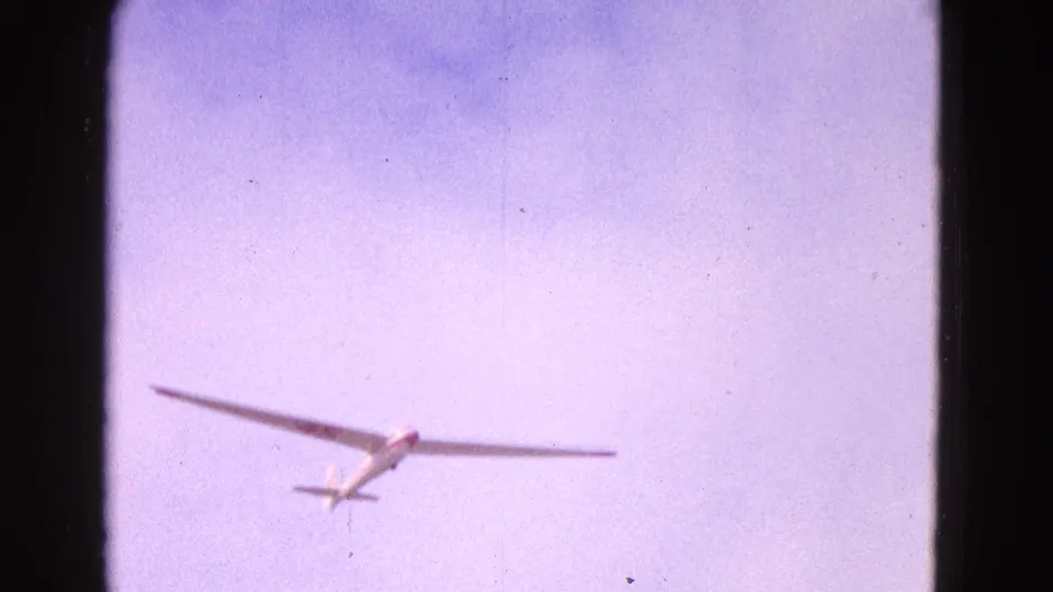 White glider with long wings and visible tail approaches across a pale sky, recorded on grainy 8mm home-movie film in 1964.