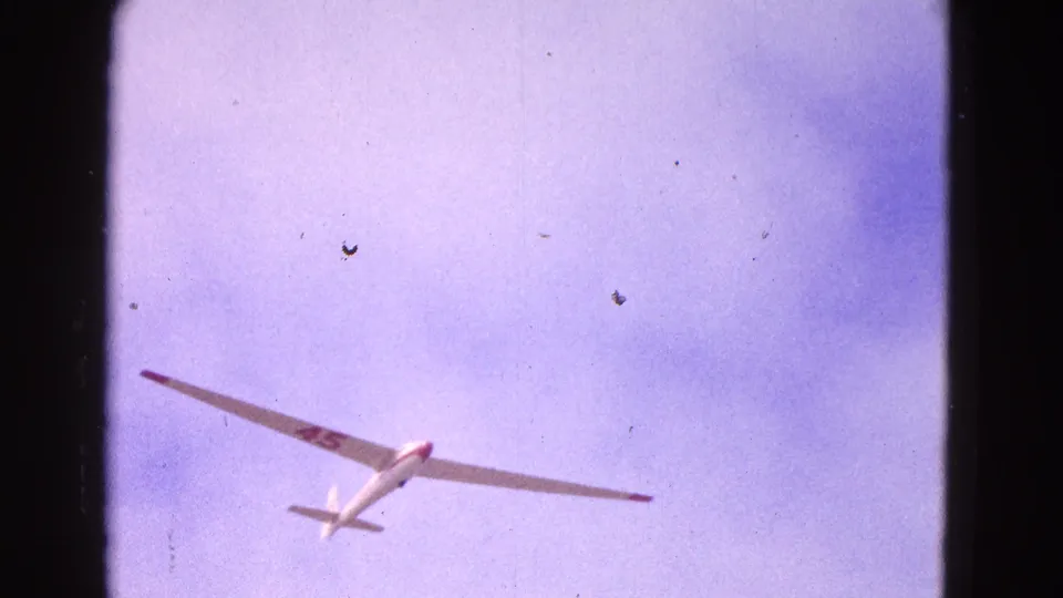 1964 home-movie frame showing a white glider crossing a blue-pink sky, with dark numbers or markings visible on the wing.