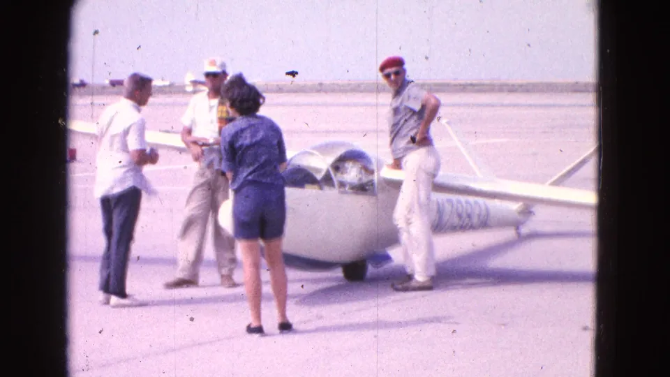 At a sunny airfield, several adults in 1960s clothing stand around a white glider with its cockpit canopy open.