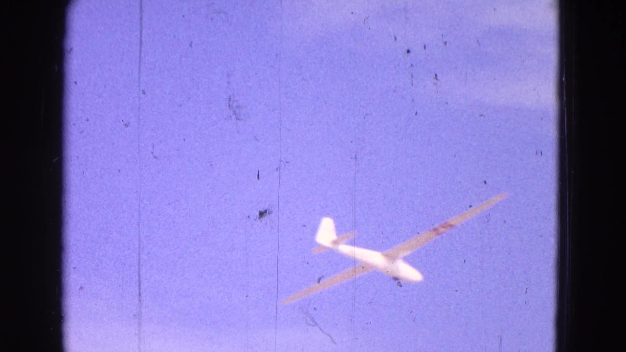 A white glider with long wings flies across a pale blue sky in the 1964 airfield footage.