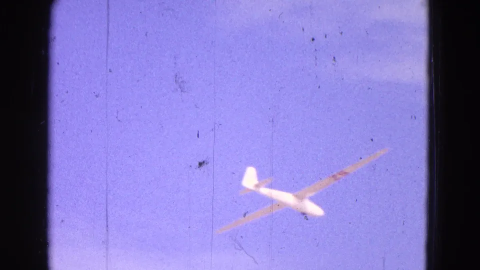 A white glider with long wings flies across a pale blue sky in the 1964 airfield footage.