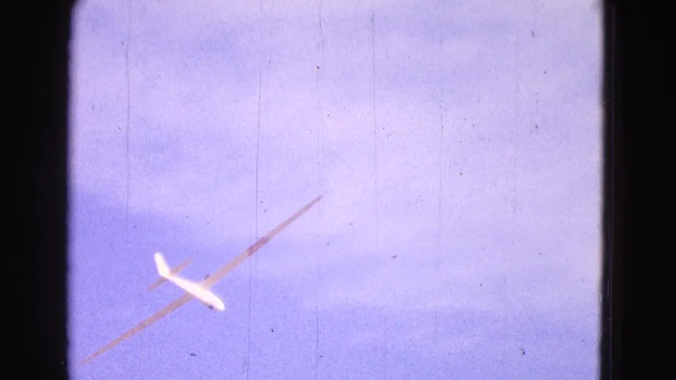 The glider is filmed from below while turning through pale sky, with visible 8mm dust and scratches.