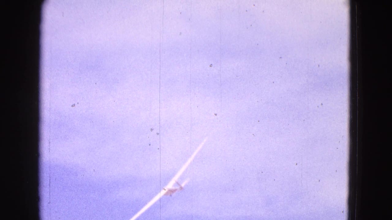 1964 8mm frame showing a distant white glider banking across a blue sky, with visible film grain, dust, and dark gate edges.