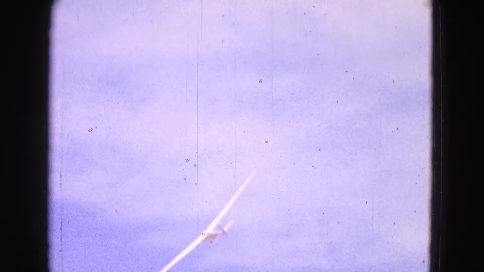 1964 8mm frame showing a distant white glider banking across a blue sky, with visible film grain, dust, and dark gate edges.