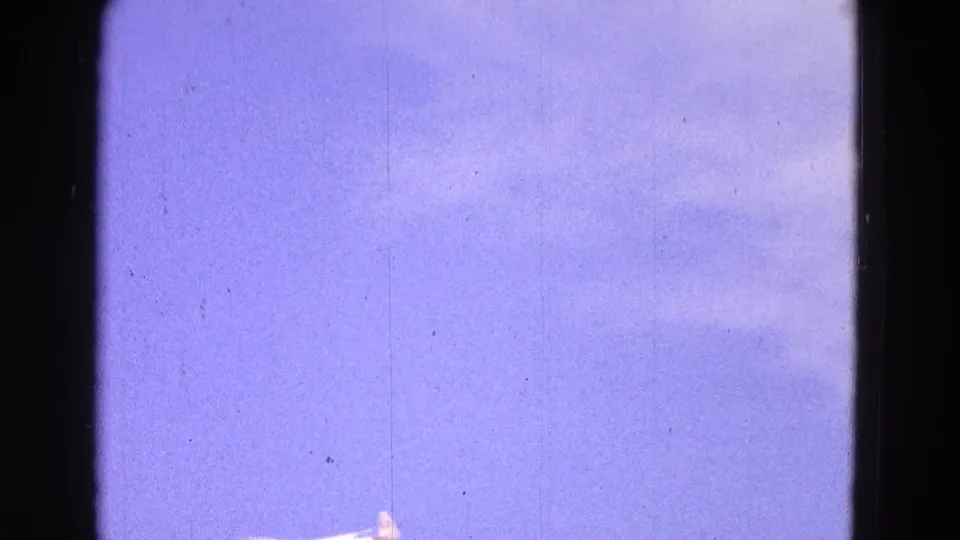 A distant white glider appears low in the 1964 8mm frame, set against blue sky and faint clouds with dust and scratch marks.