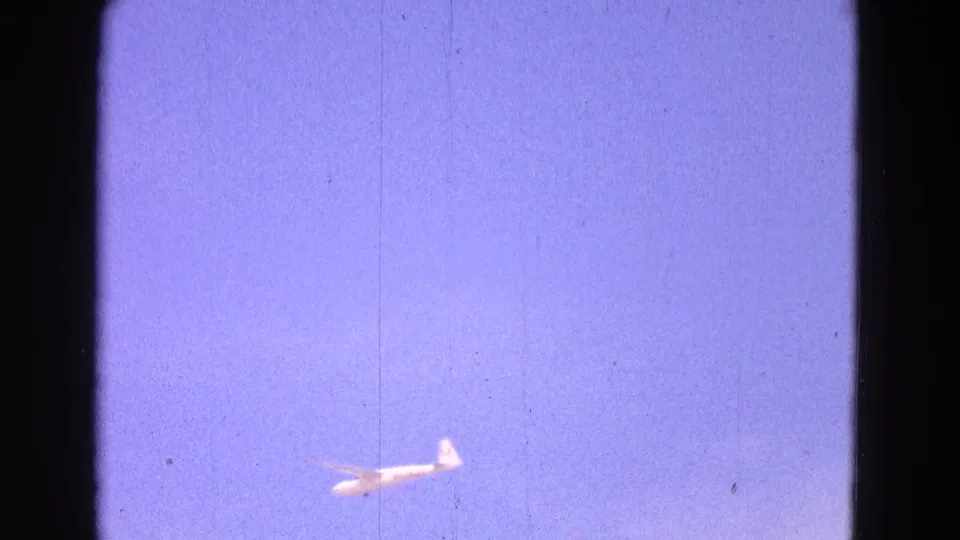 1964 8mm frame of a small white glider moving through the lower sky, with soft focus, grain, scratches, and a blue-purple tint.