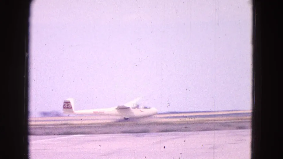 A white glider with a visible tail marking moves across an airfield runway under a pale sky, with faded 8mm color and scratches.
