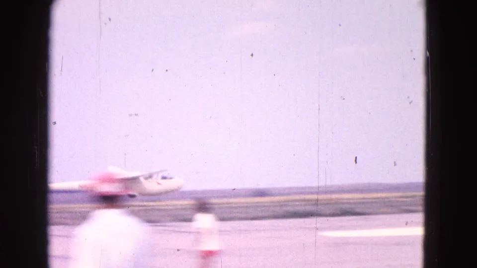 A blurred foreground figure crosses the home-movie frame while the small white glider continues along the runway in the background.