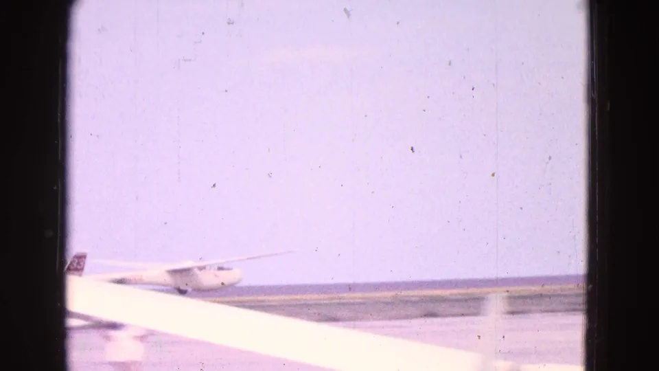 The glider is seen farther across the runway, partly blocked by a bright foreground object and framed by faded 8mm film edges.