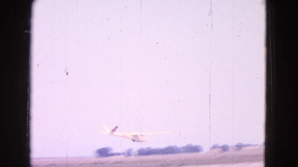1964 home-movie frame showing a small white glider low near a rural airfield outside Washington, DC, with soft focus and film borders.
