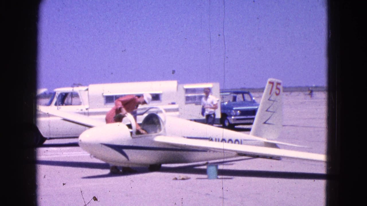 Man in red shirt climbs into a white glider on a sunny tarmac with vehicles behind him.