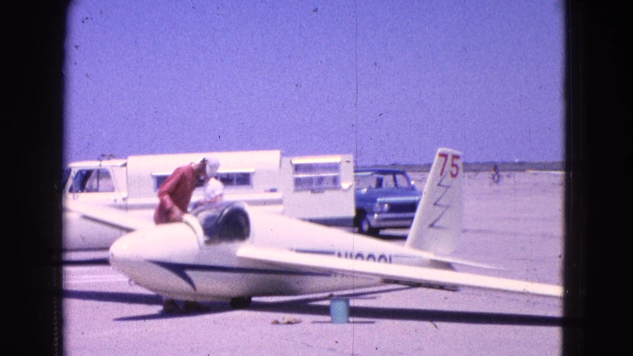 A man in a red shirt and white cap leans into the cockpit of a white glider on a Washington DC tarmac, with vehicles behind.
