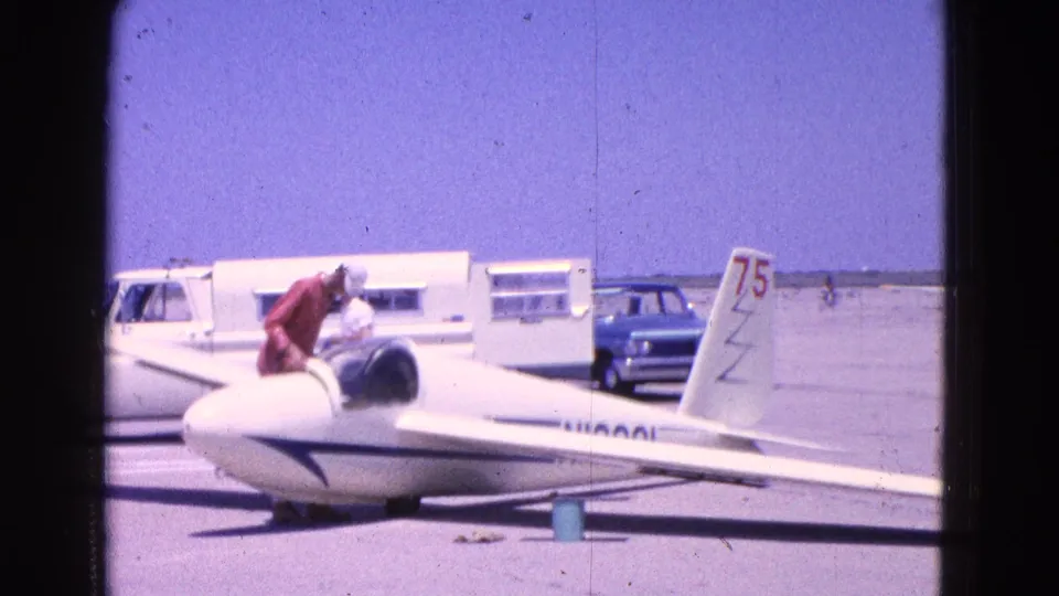 A man in a red shirt and white cap leans into the cockpit of a white glider on a Washington DC tarmac, with vehicles behind.