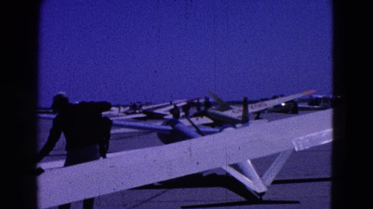 Man beside a white glider wing on an airfield with other gliders in the background.