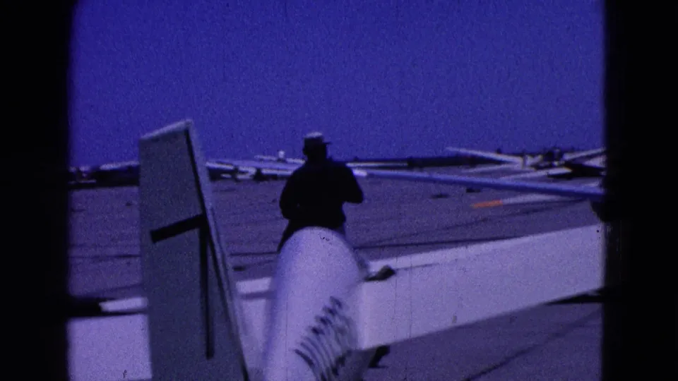 1964 Washington DC home-movie frame with a man near a glider tail and wing, seen against an open paved airfield and blue sky.