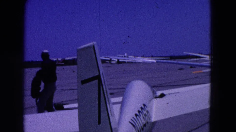 Washington DC 1964 8mm frame showing a man walking beside a glider tail and wing on an airfield with parked gliders beyond.