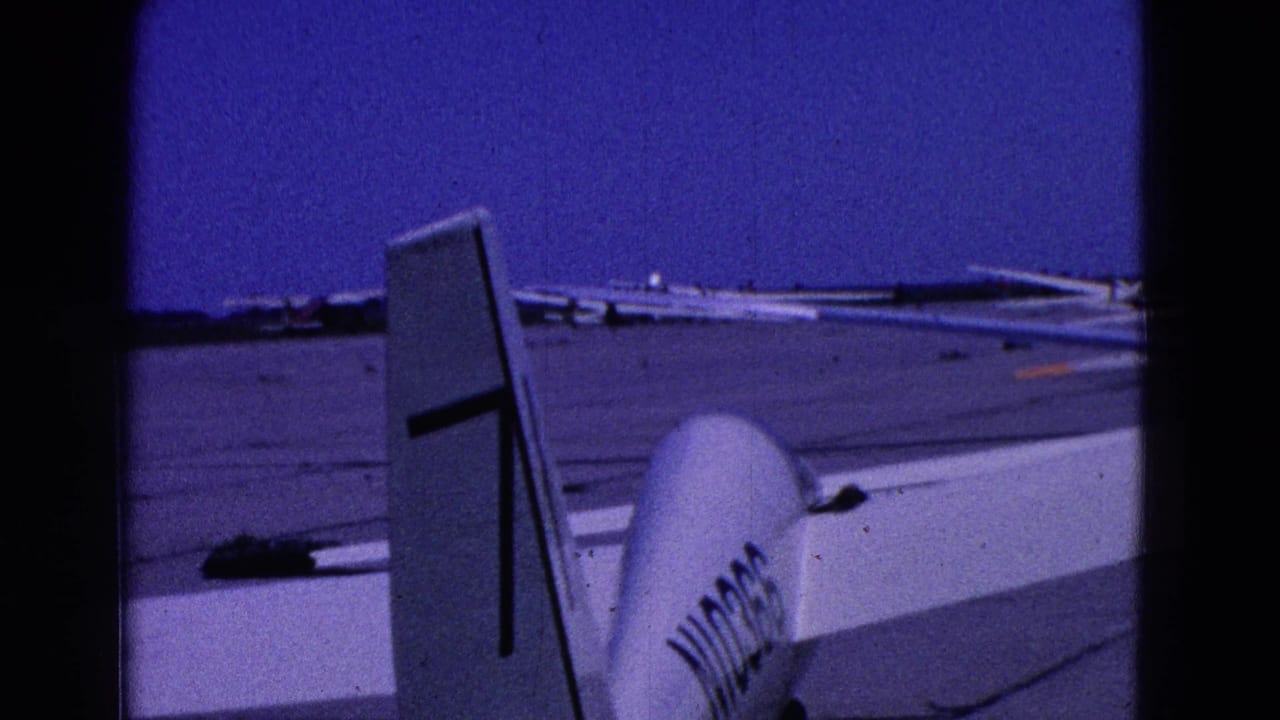 Tail and fuselage of a small white aircraft on a sunlit tarmac with other planes in the distance.