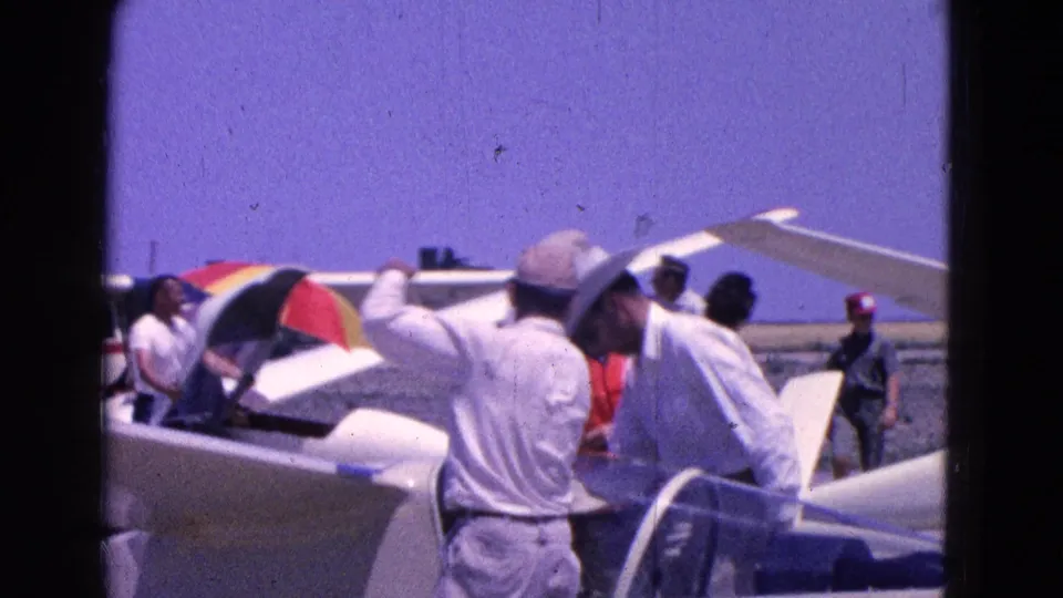 1964 Washington, DC home-movie frame showing men in light shirts around a small aircraft, with an open canopy and striped umbrella.