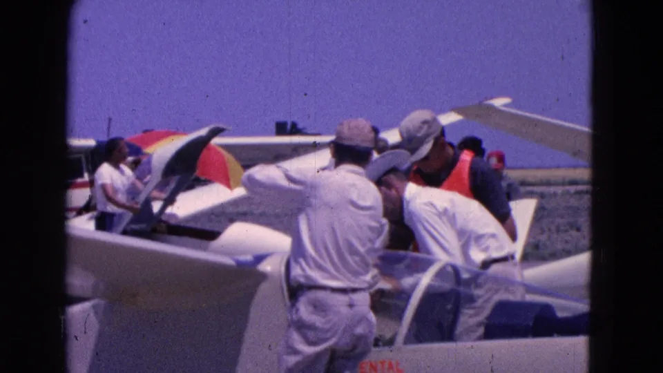 1964 Washington, DC frame showing men preparing or inspecting a small aircraft cockpit area beside an open canopy on the tarmac.