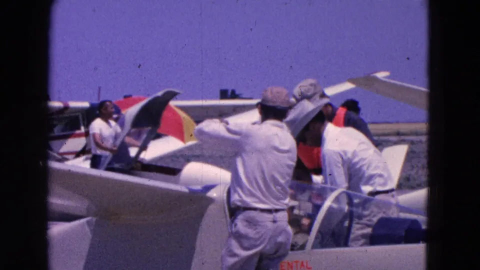 1964 Washington DC home-movie frame showing men in hats and light shirts working around white gliders at an airfield.