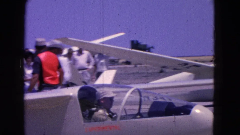 A 1964 home-movie frame shows a pilot seated in a white glider marked Experimental as men stand nearby at the airfield.