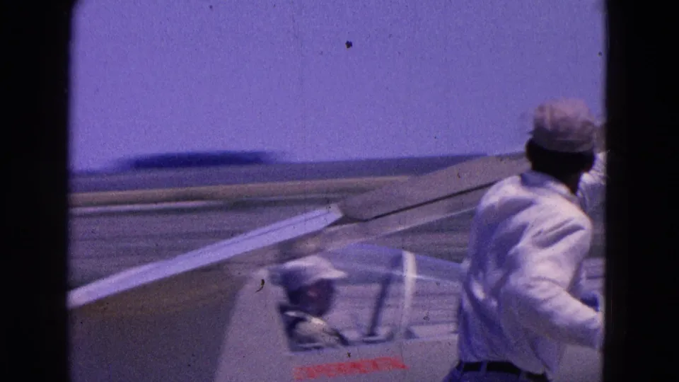 A man in a light shirt and cap runs near the wing of the small experimental aircraft on a runway.