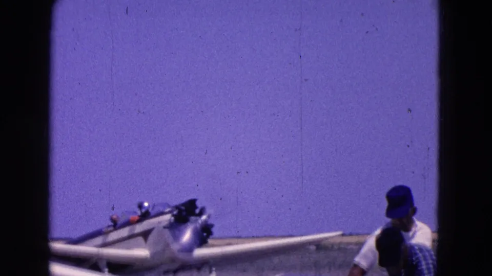 Small aircraft on a dusty field with a man in a blue cap nearby, recorded in 1964 8mm home-movie footage from Washington, DC.