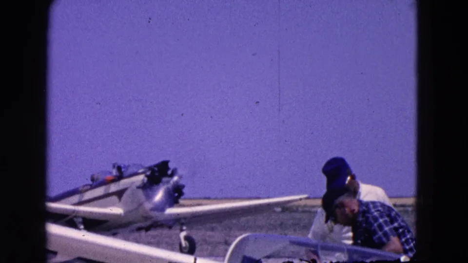 1964 home-movie frame shows two adults near a small single-engine aircraft on an open airfield, with one person by the cockpit.