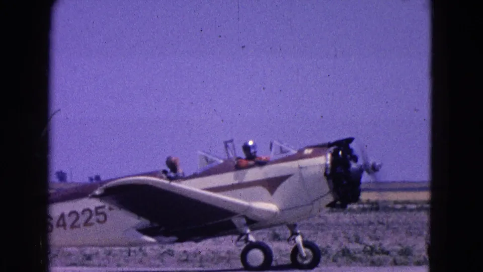 1964 8mm frame shows a close side view of the small aircraft on the airfield, with cockpit, wing root, and landing gear visible.