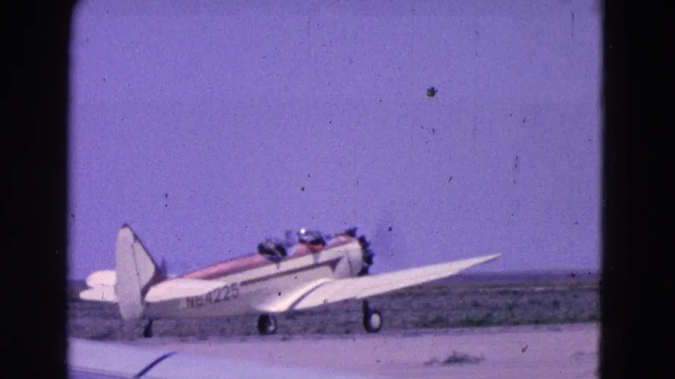 A wider 1964 home-movie view shows the small aircraft in profile on an open airfield with sky and low ground behind it.