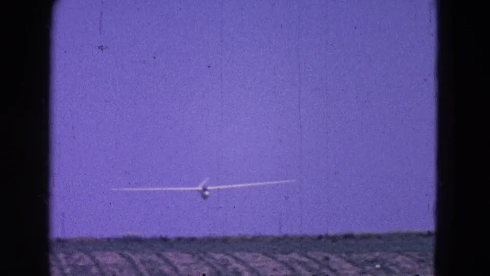 A glider approaches or passes in the distance over flat fields, with strong 8mm grain and purple color shift visible in the frame.