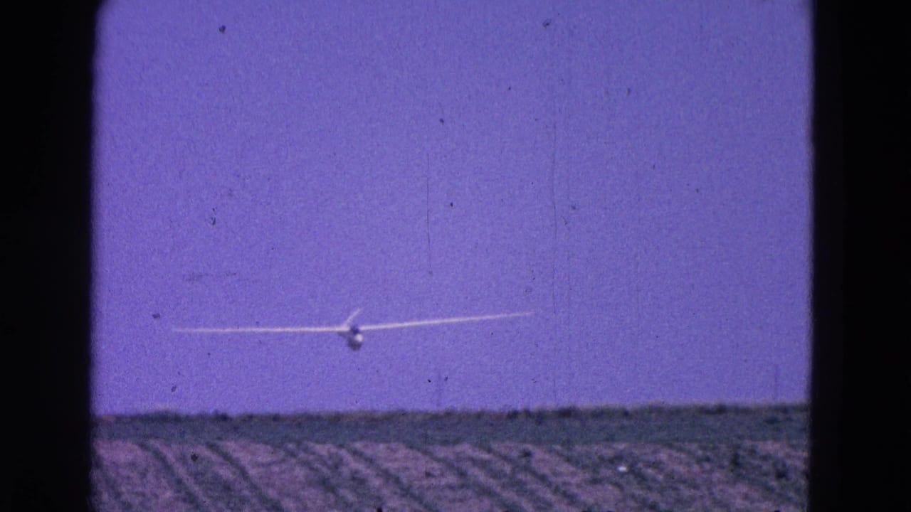 White glider flying low over plowed fields beneath a purple-blue sky.