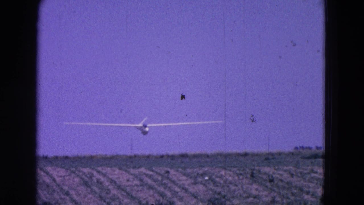 The white glider remains centered over cultivated fields, framed by dark 8mm film edges and grain.