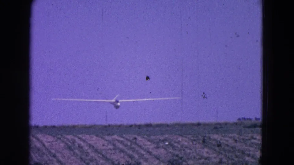 The white glider remains centered over cultivated fields, framed by dark 8mm film edges and grain.