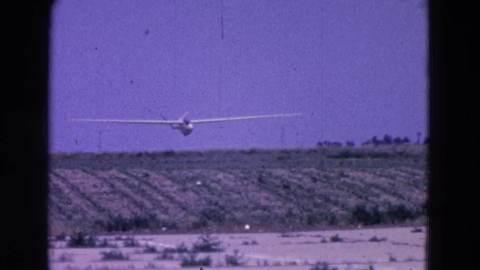 A white glider passes above rural fields and a pale open foreground area in 1960s 8mm footage.
