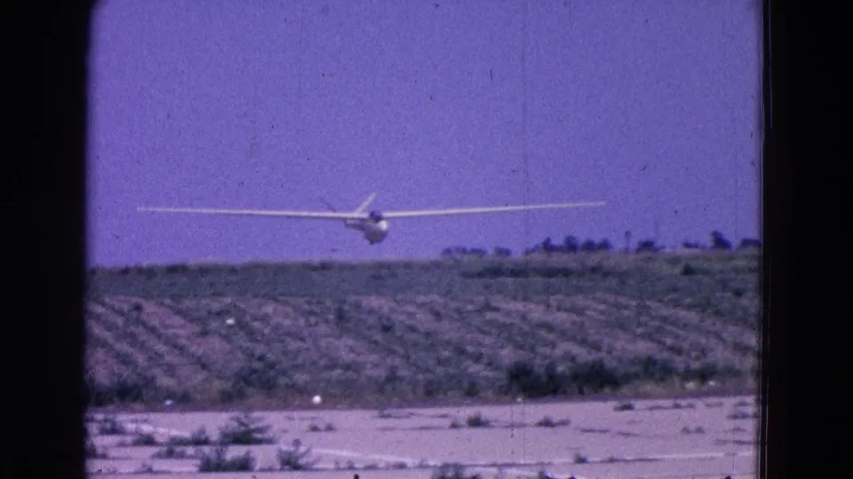 White glider crosses over cultivated land and a distant tree line in Washington DC-area 1964 home-movie footage.