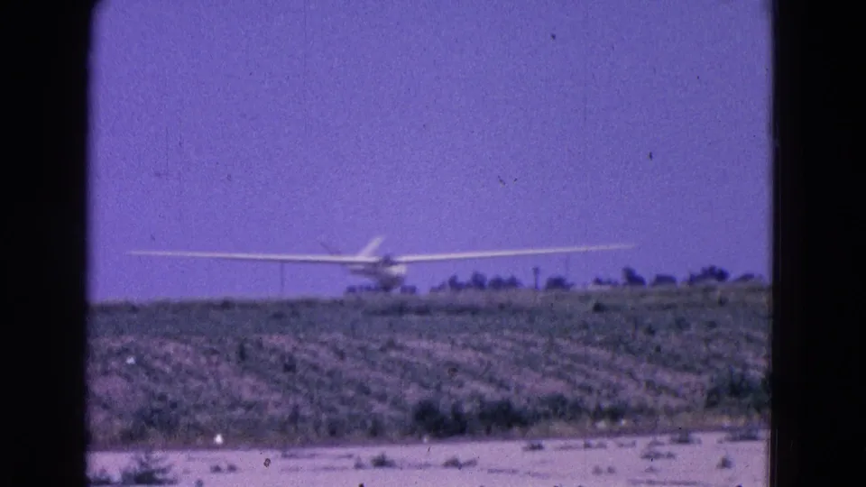 1964 8mm frame of a distant glider lined up over cultivated rural ground near Washington, DC, with sparse trees on the horizon.